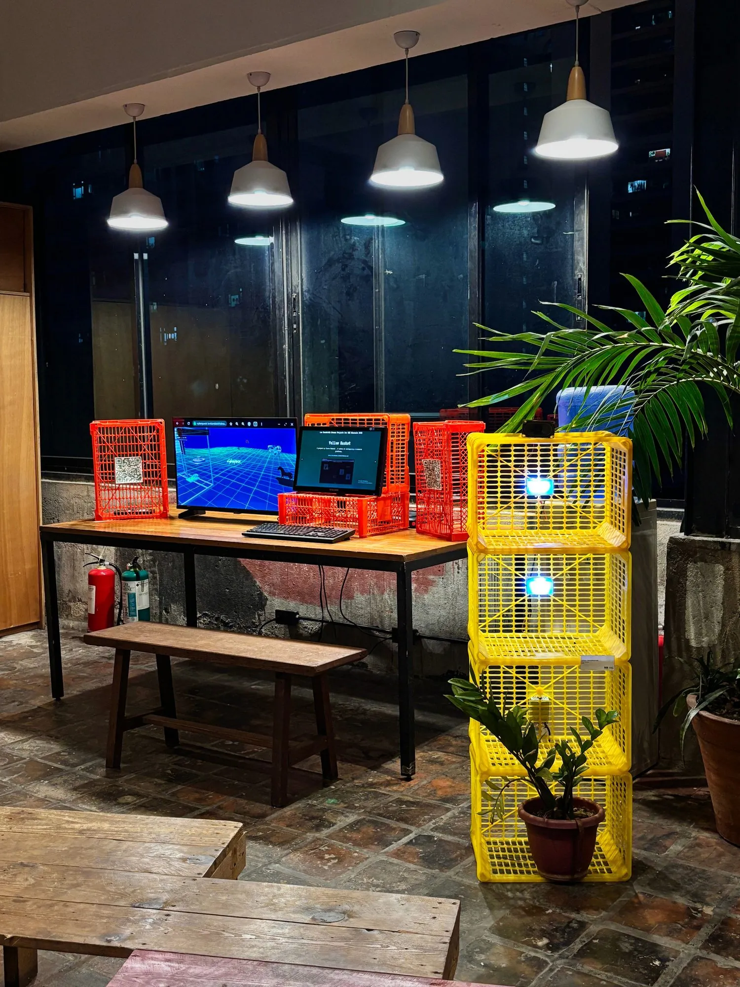 A tower of yellow plastic crates with glowing modules beside a table of screens