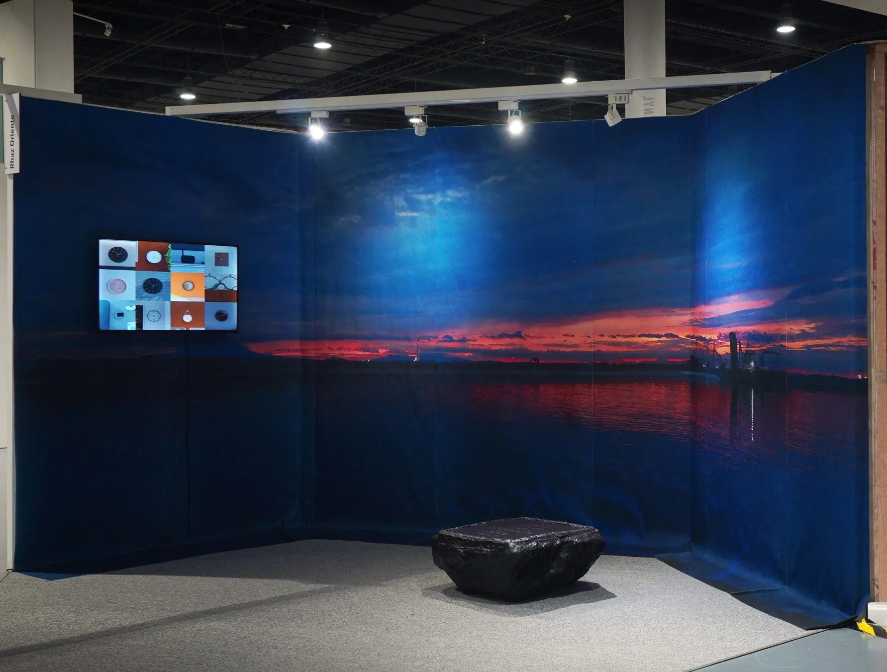 Installation view: a sunset over Manila Bay printed on fabric panels wrapped around a corner, with a screen of clocks and a black resin form on the floor