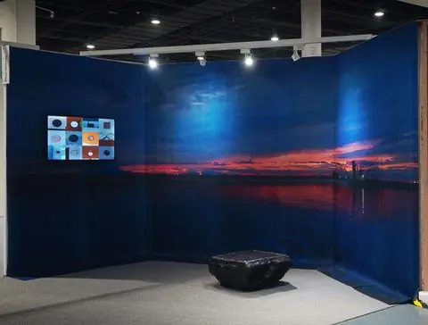 Installation view: a sunset over Manila Bay printed on fabric panels wrapped around a corner, with a screen of clocks and a black resin form on the floor